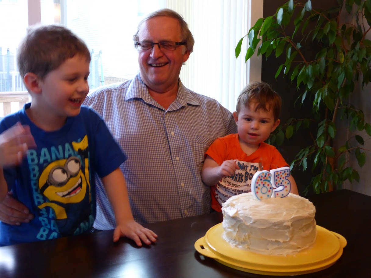 Richard on his 65th birthday with Caiden and Ethan, sitting behind a cake with candles with the number 65